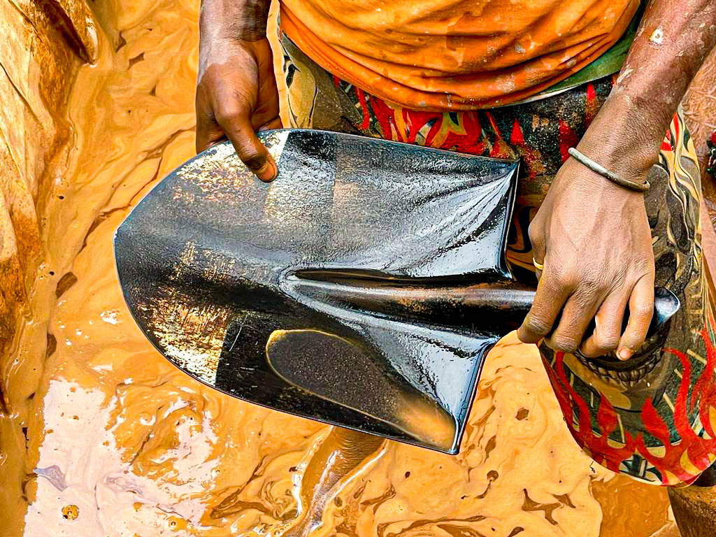 An artisanal small-scale gold miner in the Democratic Republic of Congo stands ankle-deep in water, holding a shovel which contains the glint of gold dust.